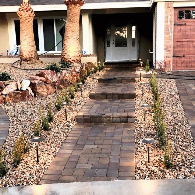 A beautifully landscaped front entrance in Las Vegas featuring a brick paver staircase illuminated by warm pathway lights, leading to a welcoming front door framed by palm trees and lush greenery