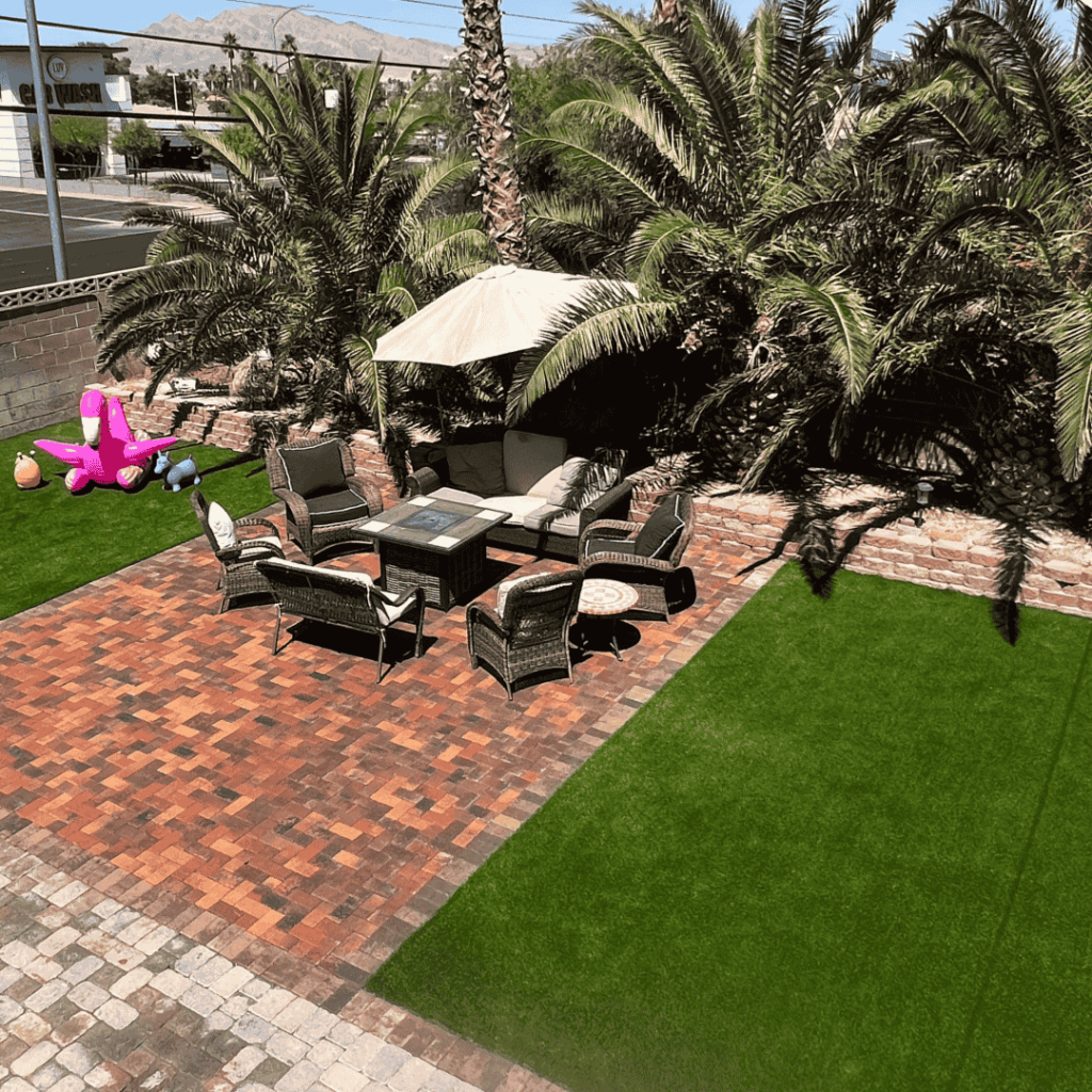 An outdoor patio with brick pavers, a fire pit seating area, artificial turf, palm trees